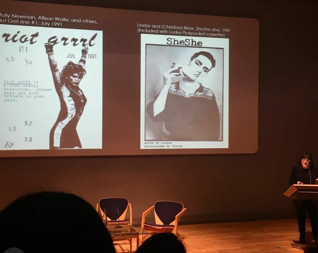 Prof. Buszek stands at a podium while lecturing at Cambridge. Two magazine covers are on the screen behind her.