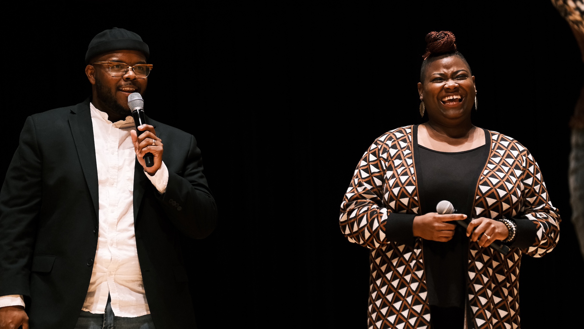 Two people standing onstage. One is talking into a mic and the other is smiling.