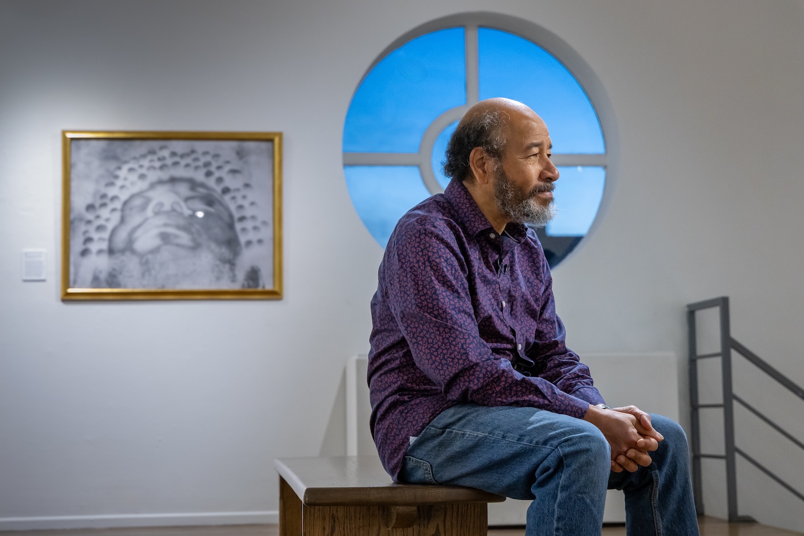 Artist Albert Chong sits on a bench in the Emmanuel Art Gallery surrounded by his artwork.