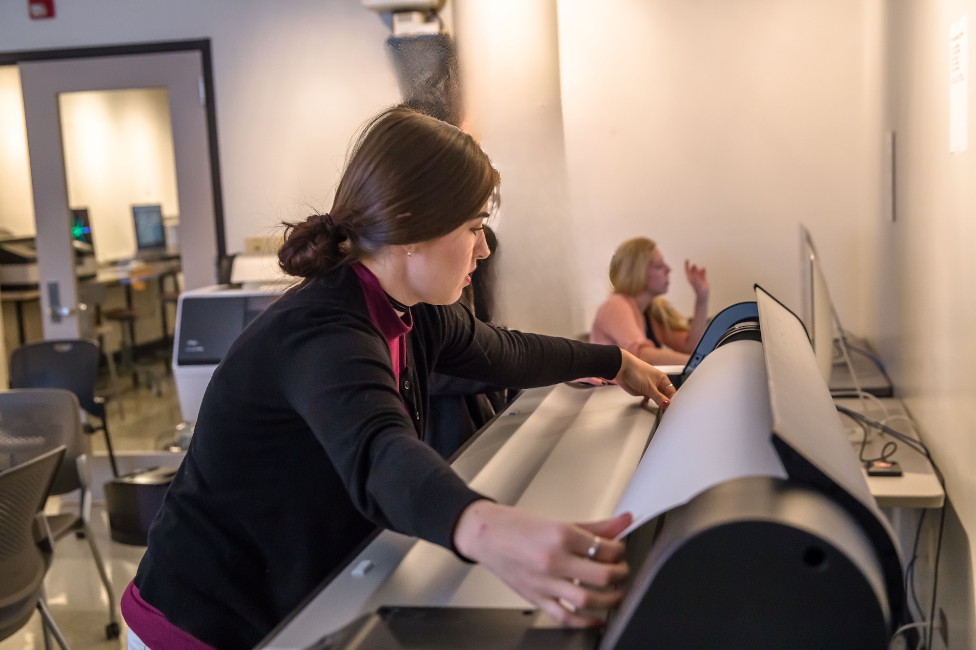 A person loads paper into an oversize printer in the foreground. A person in the background is sitting at a computer.