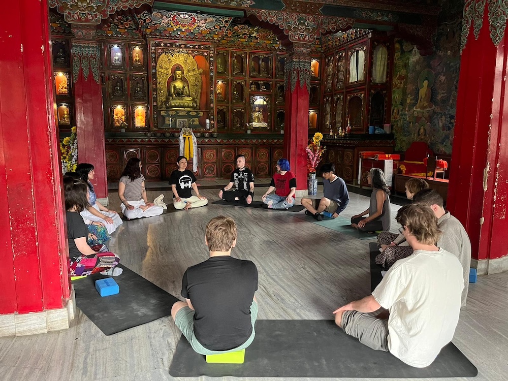 Group of 13 people sitting cross-legged in a circle practicing meditation in a buddhist temple.”
 title=